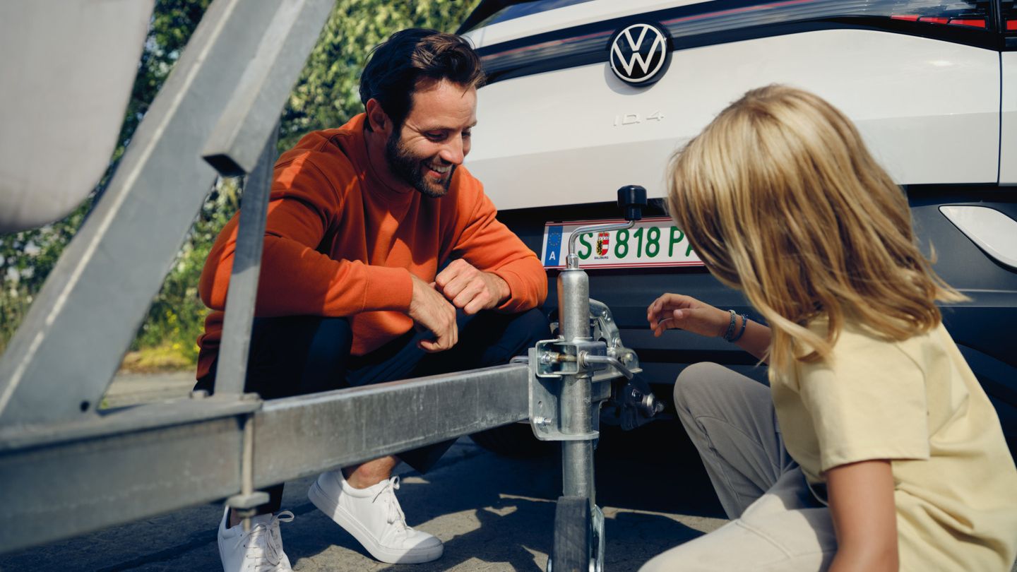 Ein Mann und ein Mädchen schauen sich die Anhängerkupplung eines VW ID.4 an. Ein Anhänger mit Boot ist daran angekoppelt.