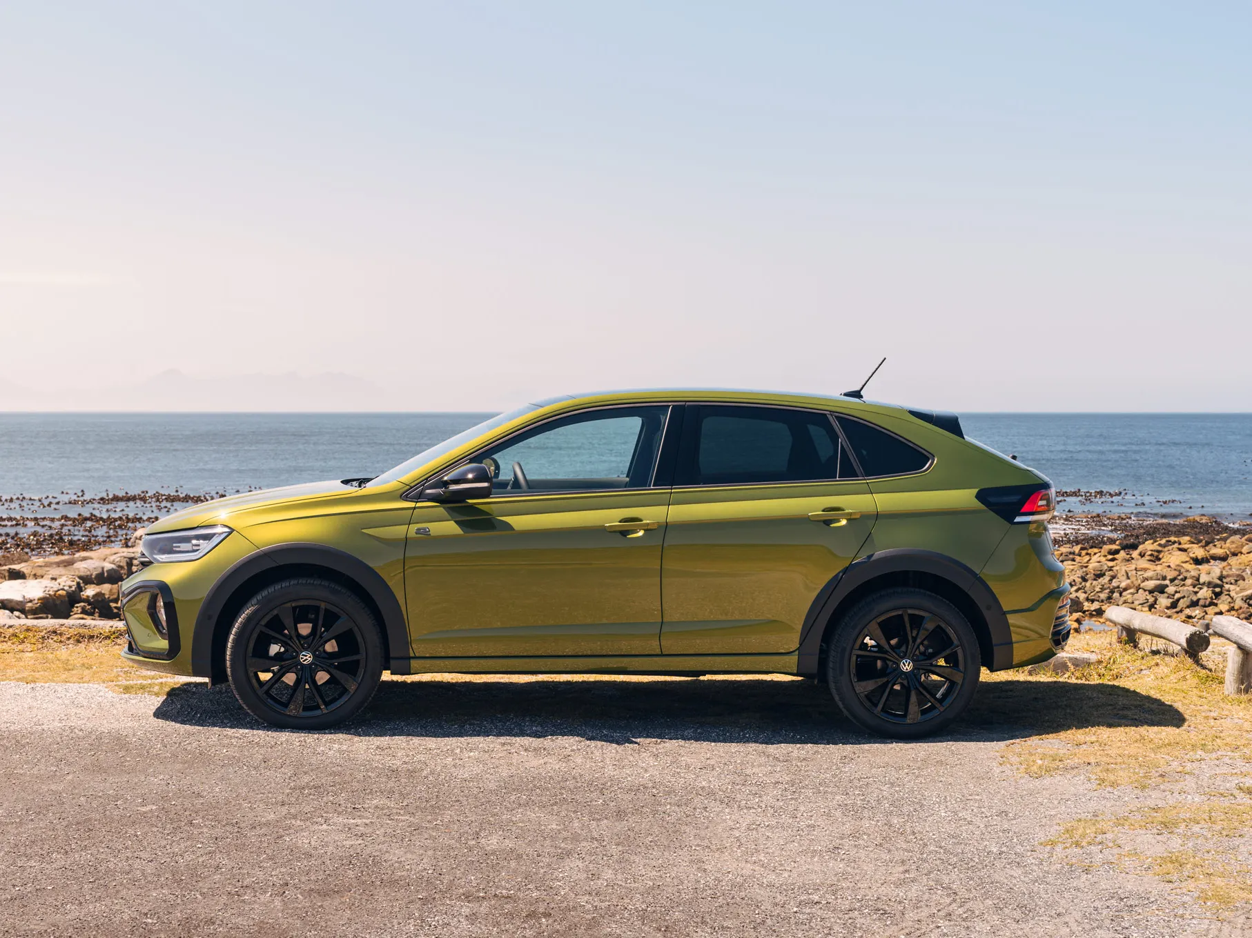 Ein grüner Taigo Black Edition steht seitlich am Meer mit sichtbaren Felsen am Ufer und einem klaren Himmel im Hintergrund. Das Auto hat schwarze Reifen und dunkle Fenster.
