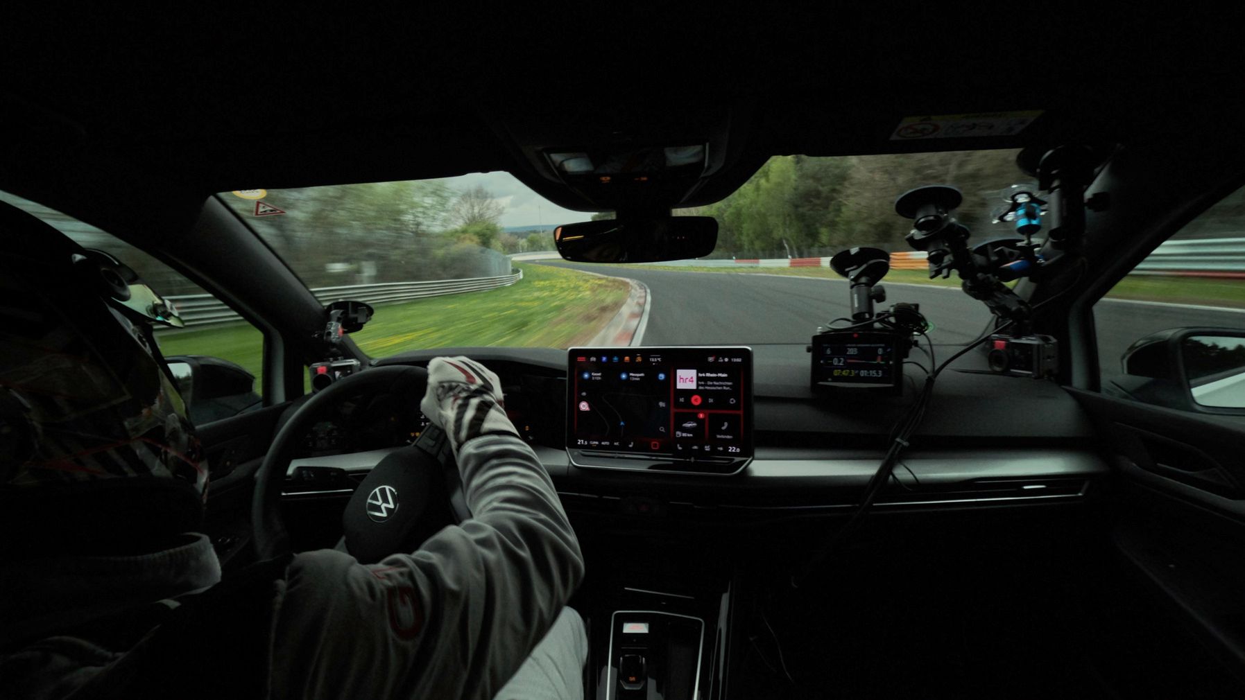 Ein Fahrer mit Helm und Handschuhen steuert ein Auto auf einer Rennstrecke. Die Perspektive zeigt den Innenraum des Fahrzeugs und die Strecke vor ihm. Auf dem Armaturenbrett sind Kameras montiert.