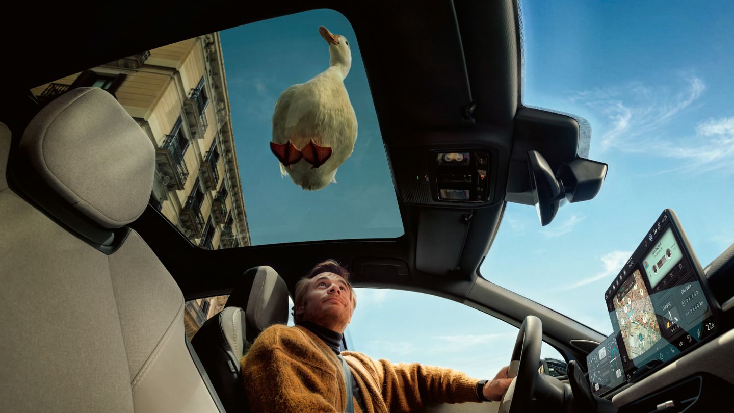 Ein Mann sitzt auf dem Fahrersitz eines VW ID. Polo und schaut nach oben durch das Panoramadach, auf dem eine weiße Ente sitzt.