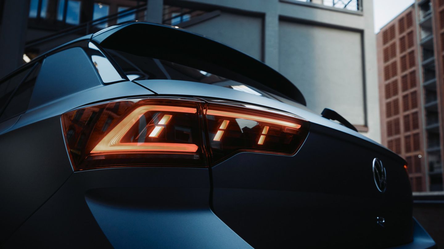 Nahaufnahme des Rücklichts eines modernen Autos, möglicherweise eines Volkswagen. Das Fahrzeug steht vor einem städtischen Hintergrund. Die Beleuchtung der Rücklichter ist eingeschaltet.