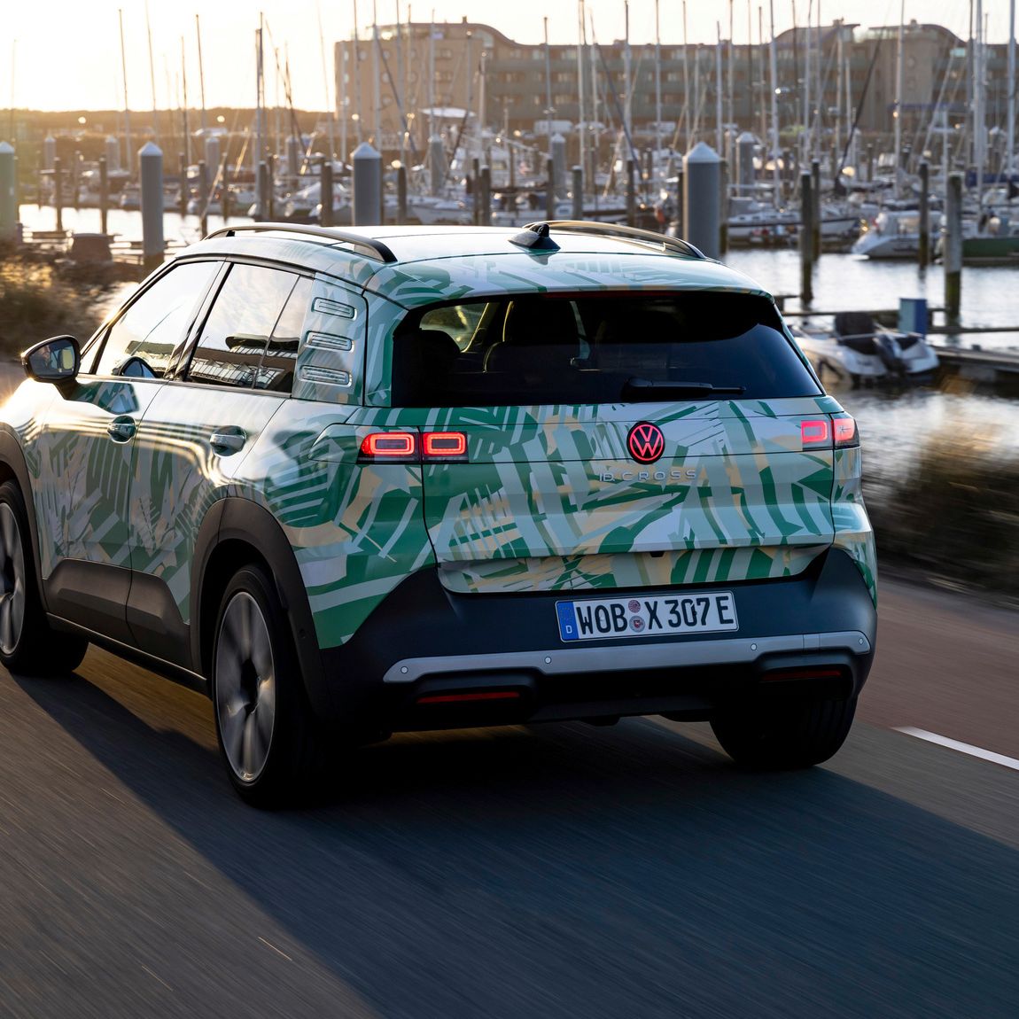 Ein Volkswagen-Elektroauto fährt auf einer Straße entlang eines Hafens bei Sonnenuntergang. Im Hintergrund sind viele Segelboote und Yachten zu sehen. Die Straße und die Landschaft wirken ruhig und friedlich.