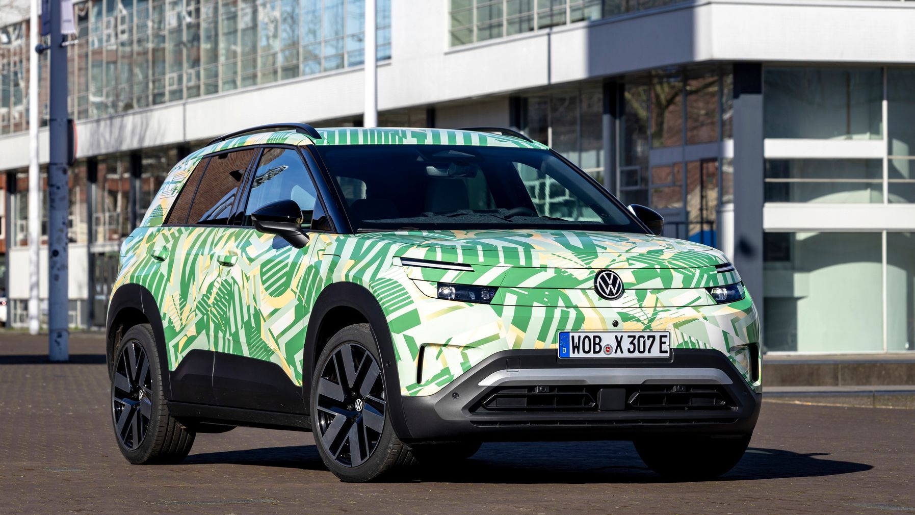 Ein Volkswagen Elektro-SUV steht vor modernen Gebäuden. Das farbenfrohe Fahrzeug hat ein grünes und cremefarbenes Camouflage-Muster mit Blattmotiven. Das Nummernschild zeigt eine Zulassung aus Wolfsburg.