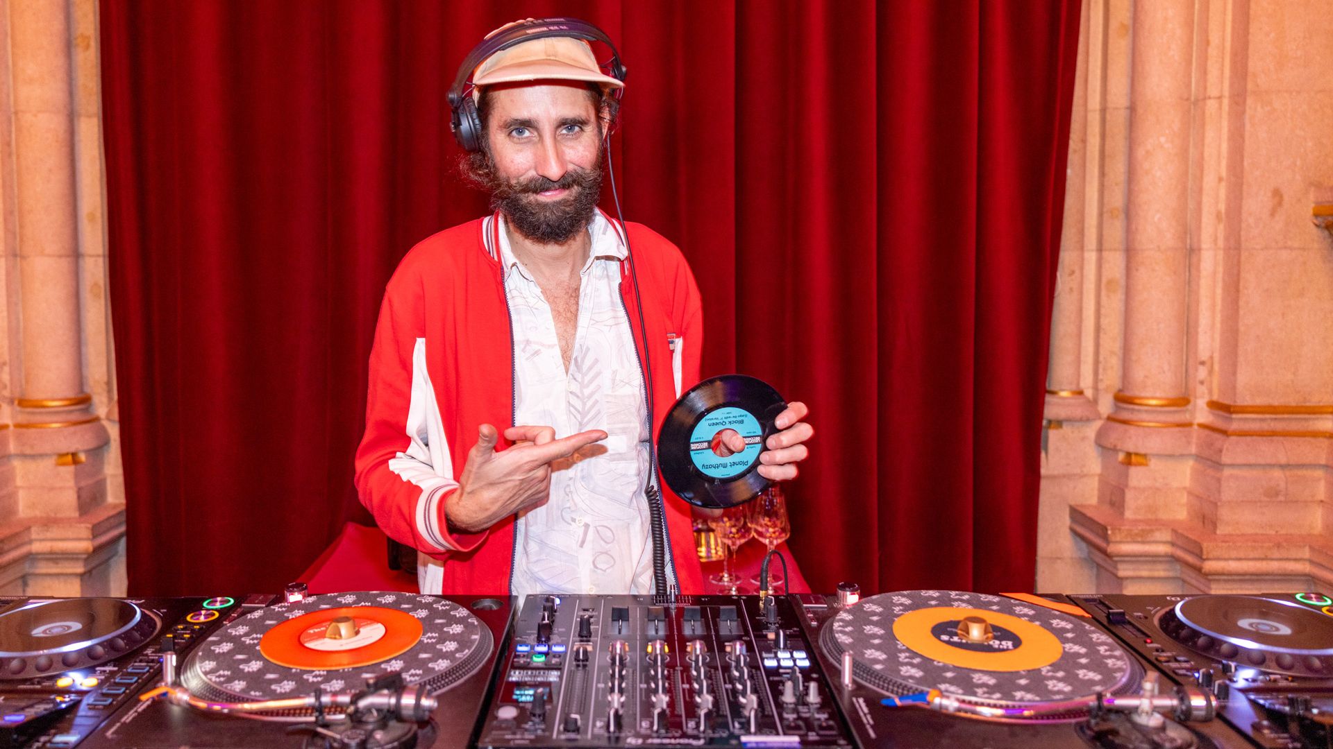 Ein DJ mit Bart und Kopfhörern steht vor einem roten Vorhang und trägt eine rote Jacke und eine beige Kappe. Er lächelt in die Kamera, hält eine kleine Schallplatte in der einen Hand und zeigt mit der anderen Hand darauf. Vor ihm steht ein großes DJ-Pult mit mehreren Mischpulten und Plattentellern.
