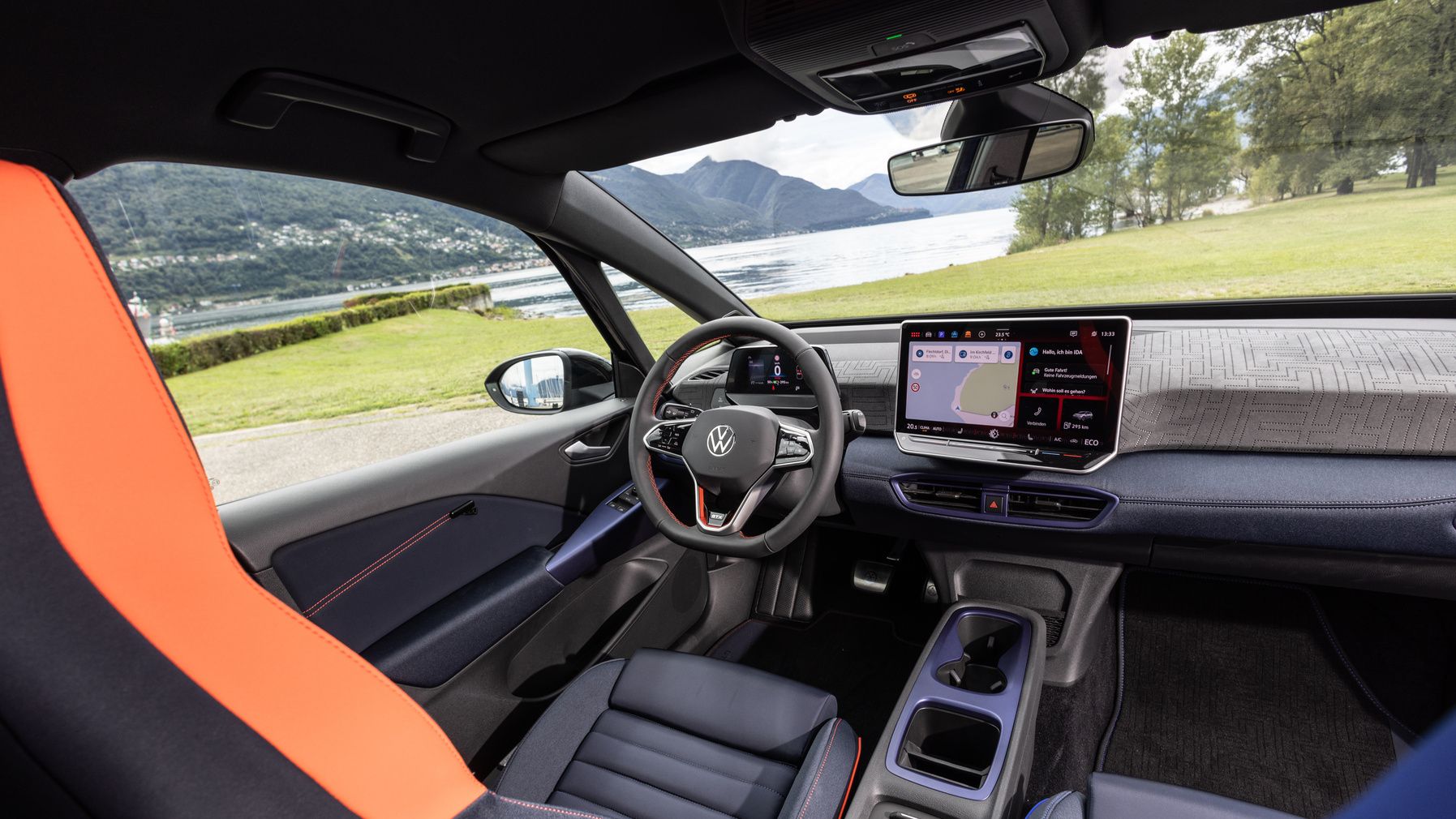 Das Bild zeigt den Innenraum eines modernen Autos mit einem digitalen Armaturenbrett und einem großen Bildschirm. Man sieht auch einen sportlichen Fahrersitz in Schwarz und Orange. Im Hintergrund befindet sich eine landschaftlich reizvolle Umgebung mit einem See und Bergen.