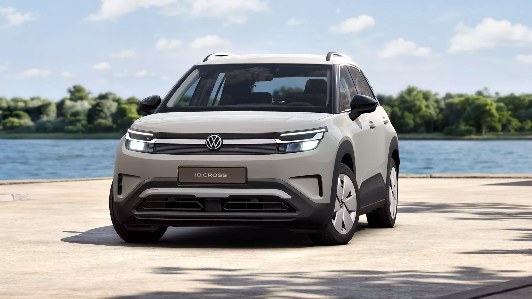 Ein heller, moderner VW ID. Cross steht auf einer Uferpromenade vor einem See, im Hintergrund Bäume und blauer Himmel mit wenigen Wolken. Das Auto ist frontal leicht schräg zu sehen, mit LED-Frontscheinwerfern und VW-Logo in der Mitte.