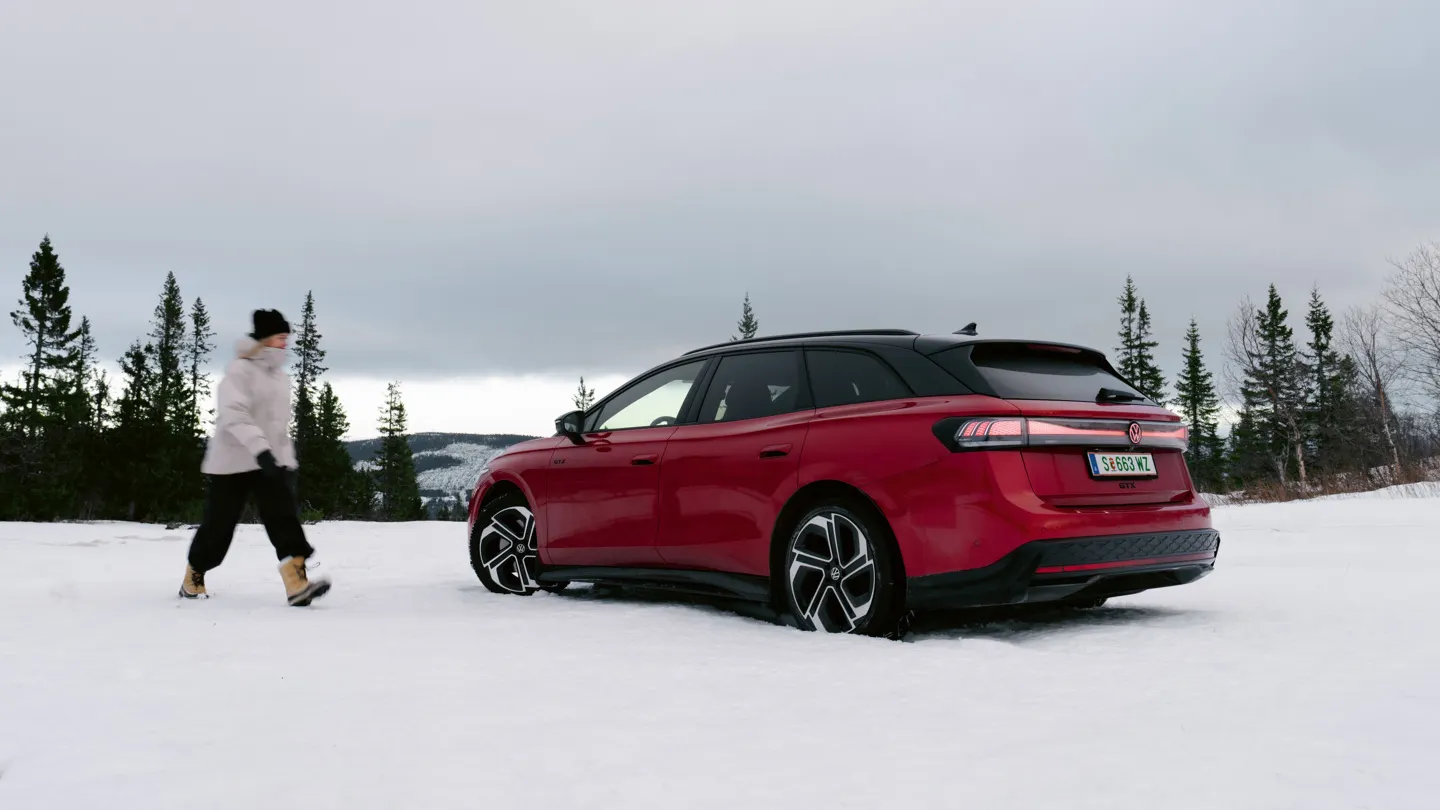 Eine Frau geht durch den Schnee auf einen roten VW ID.7 Tourer GTX zu, der in einer winterlichen Landschaft steht.