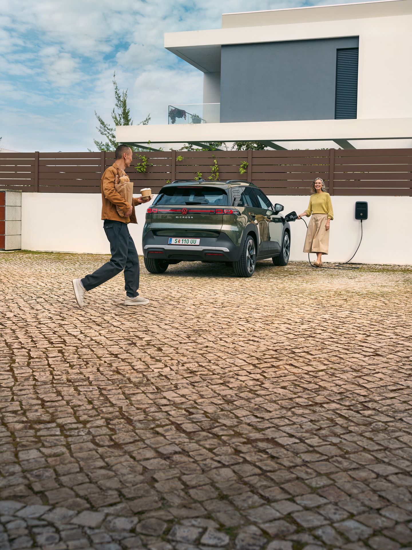 Eine Frau steckt ein Ladekabel einer Wallbox in einen grünen VW ID. Cross, ein Mann geht auf sie zu und hat Kaffeebecher in der Hand und Brot unter dem Arm.