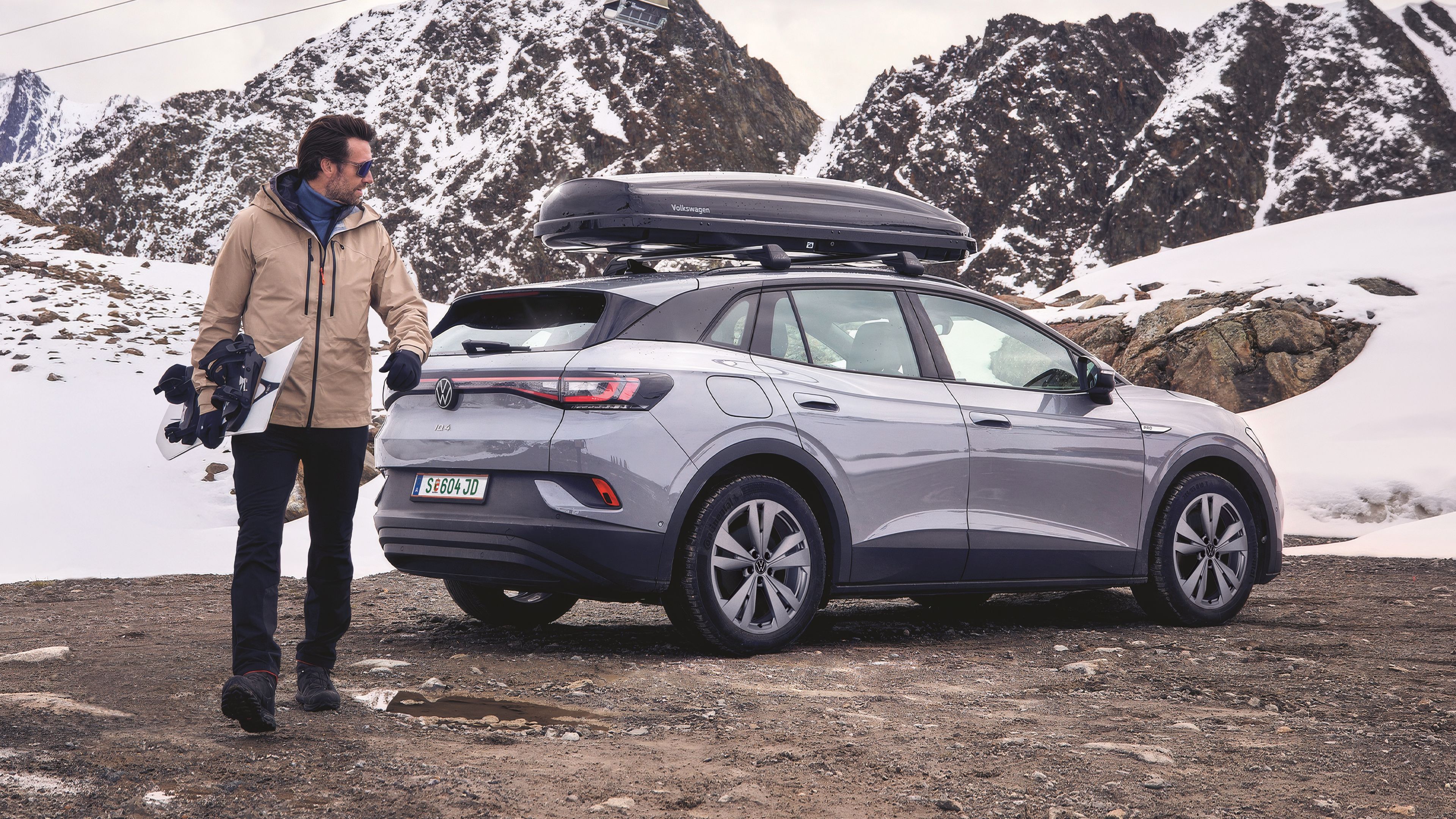 Ein Mann steht neben einem grauen Volkswagen SUV mit Dachbox. Er hält ein Snowboard in der Hand und trägt Winterkleidung, während der Boden matschig und teils von Pfützen bedeckt ist.