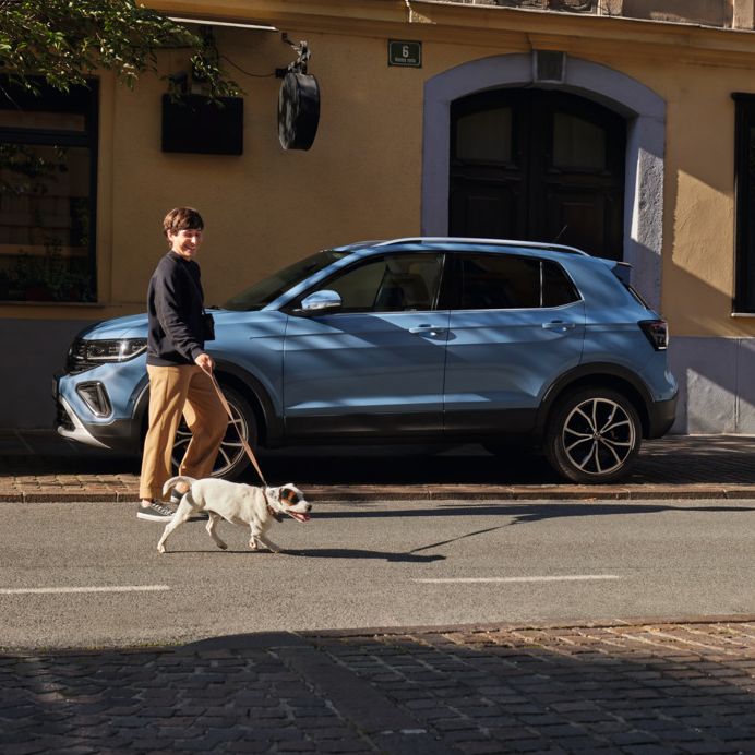 Eine Frau geht mit einem Hund an einem blauen VW T-Cross Style vorbei.