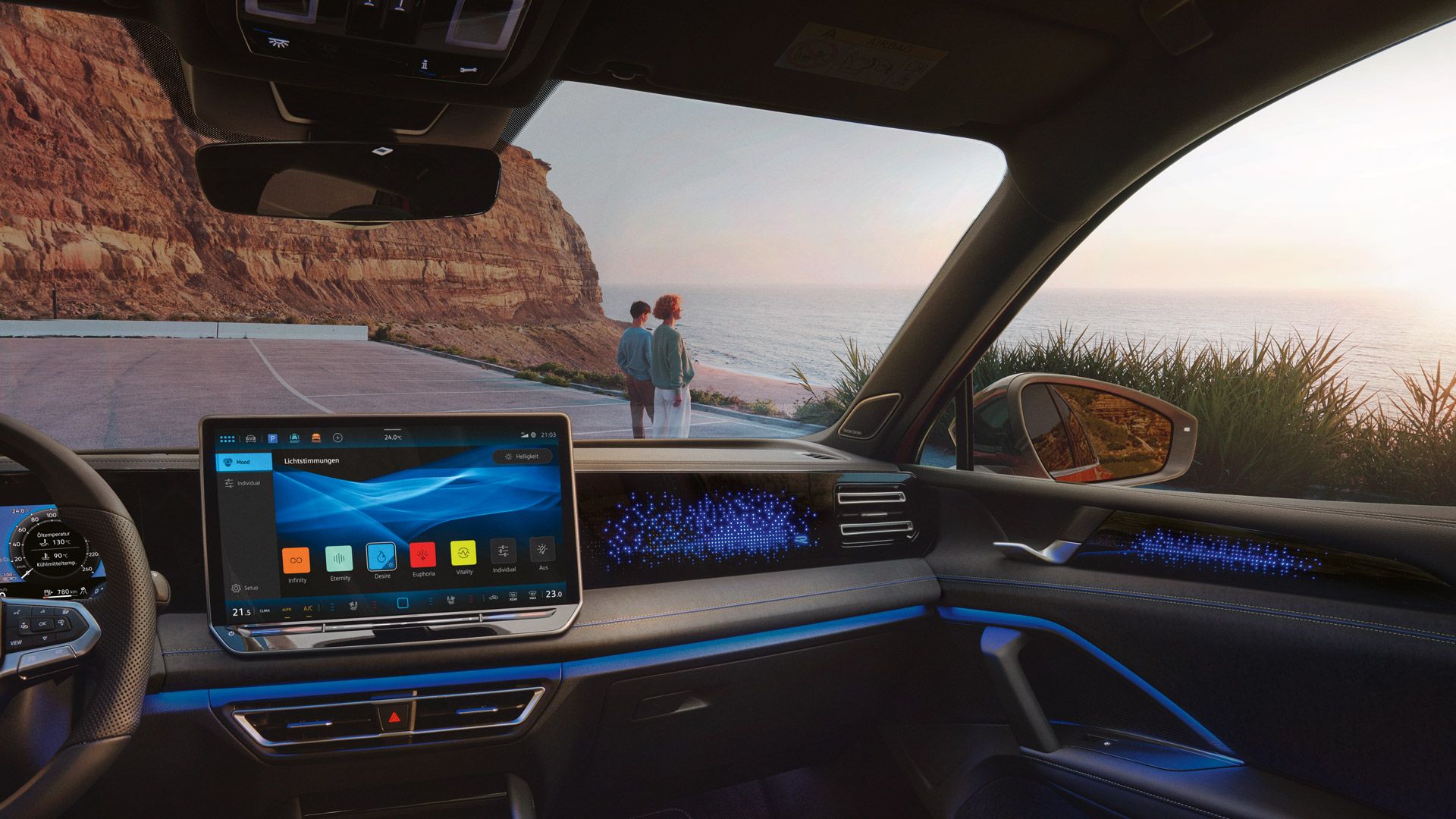 VW Tiguan mit blauer Ambientebeleuchtung. Durch die Frontscheibe ist ein Küstenlandschaft und ein auf das Meer schauendes Paar sichtbar.