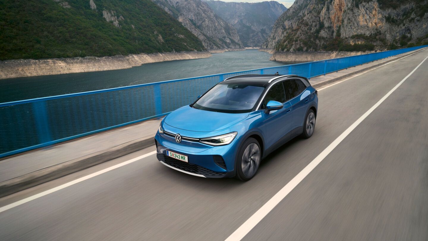 Ein blauer VW ID.4 fährt über eine Brücke, im Hintergrund ein See und Berge.