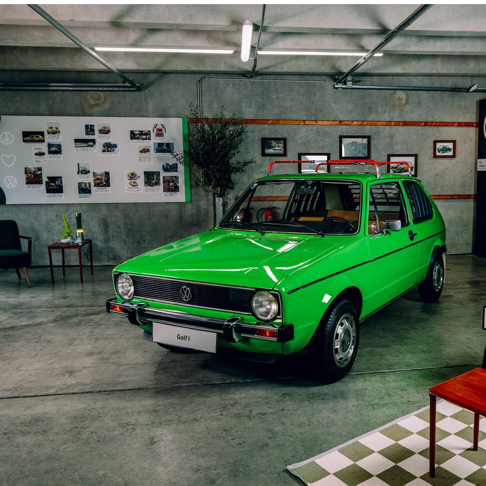 Ein grüner Volkswagen Golf 1 steht in einem Ausstellungsraum. Im Hintergrund sind Fahrzeugbilder und ein Baum zu sehen. Das Auto hat ein Retro-Kofferträgersystem auf dem Dach.