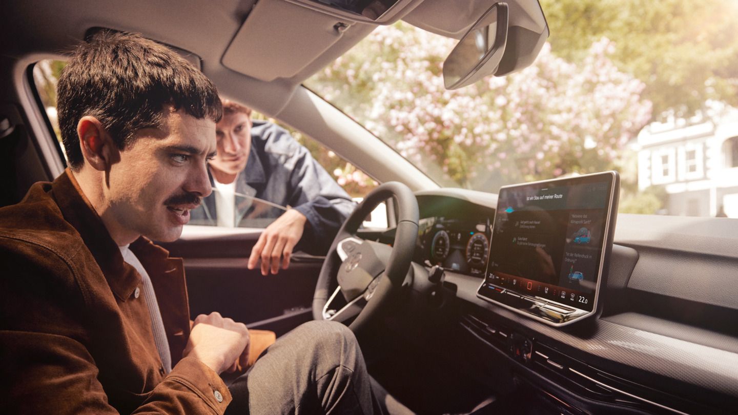 Ein Mann sitzt im neuen VW Golf und schaut auf das Infotainment-Display, ein anderer schaut durch das Fenster.