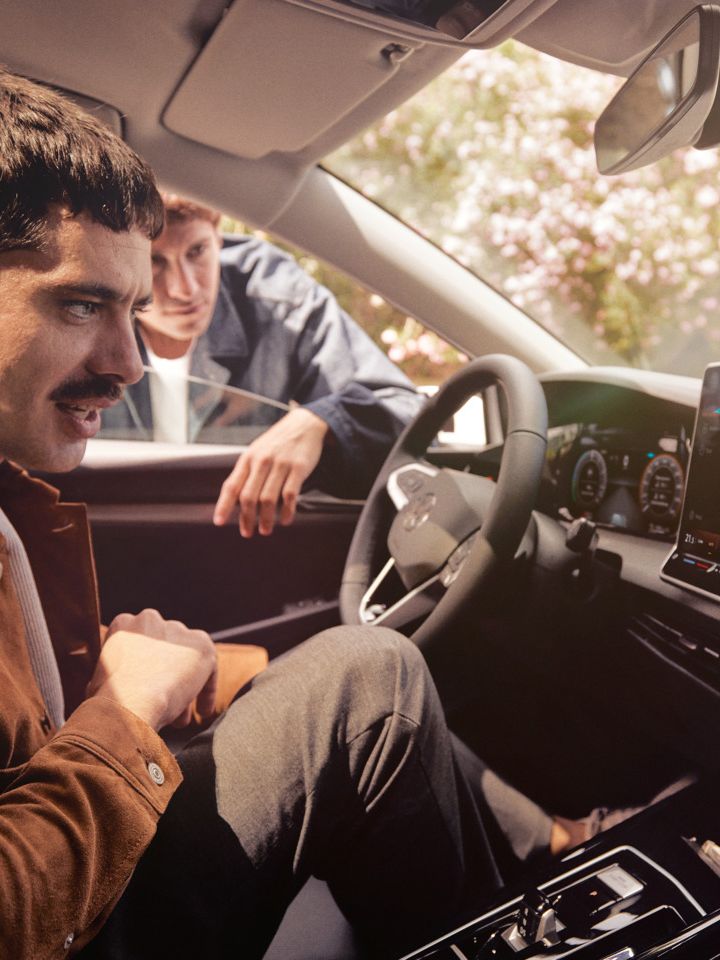 Ein Mann sitzt im neuen VW Golf und schaut auf das Infotainment-Display, ein anderer schaut durch das Fenster.
