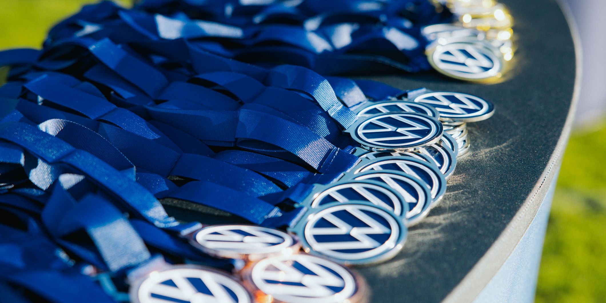 Nahaufnahme eines Tisches, auf dem zahlreiche Medaillen mit blauem Band ordentlich ausgebreitet liegen. Auf den runden Medaillen ist ein stilisiertes „VW“-Logo zu sehen. Das Bild vermittelt eine festliche oder sportliche Siegerehrungssituation.