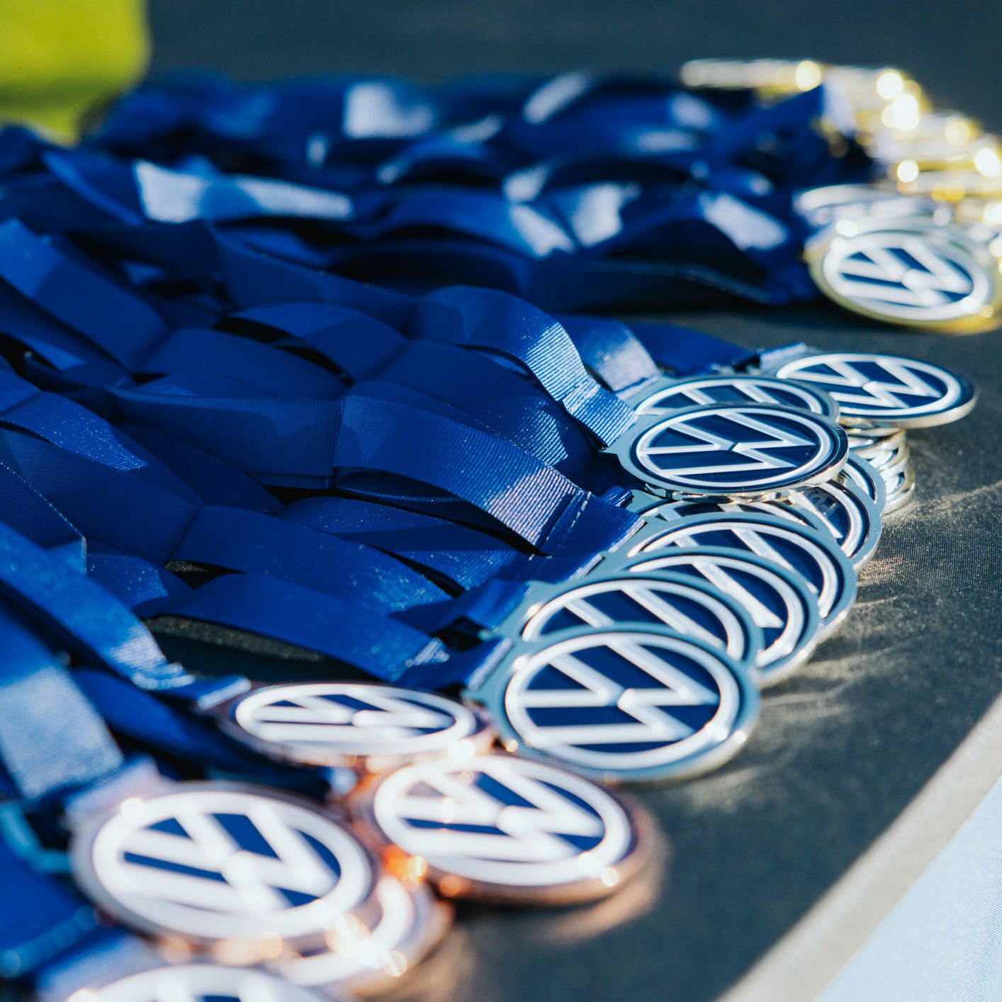 Nahaufnahme eines Tisches, auf dem zahlreiche Medaillen mit blauem Band ordentlich ausgebreitet liegen. Auf den runden Medaillen ist ein stilisiertes „VW“-Logo zu sehen. Das Bild vermittelt eine festliche oder sportliche Siegerehrungssituation.