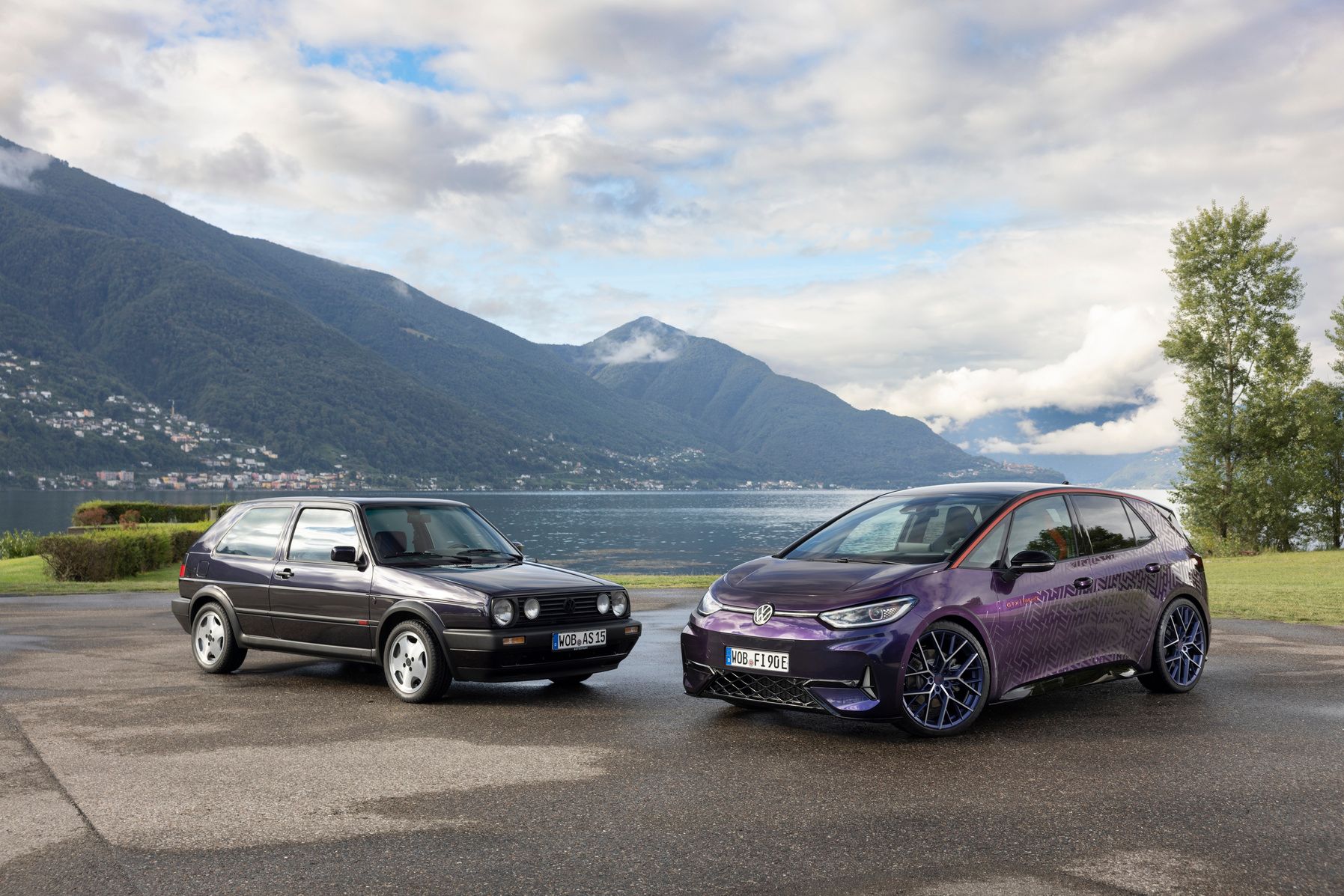 Zwei Autos stehen nebeneinander vor einer malerischen Kulisse mit Bergen und einem See im Hintergrund. Links steht ein schwarzer VW Golf aus älterer Zeit, rechts ein moderner violetter Elektroauto VW ID.4. Beide Autos sind auf einem nassen Asphalt geparkt.