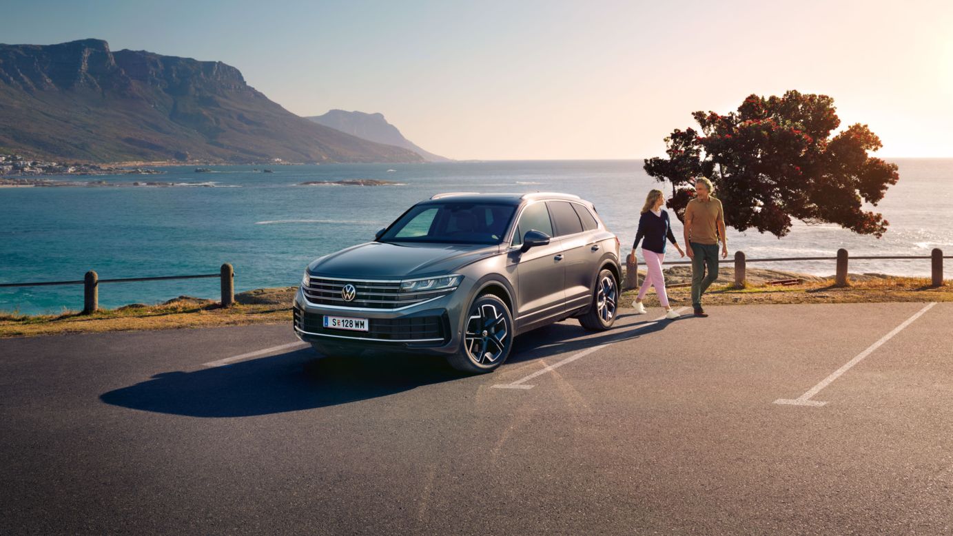 Der neue Volkswagen Touareg auf einem Parkplatz an der Küste, an ihm gehen eine Frau und ein Mann vorbei