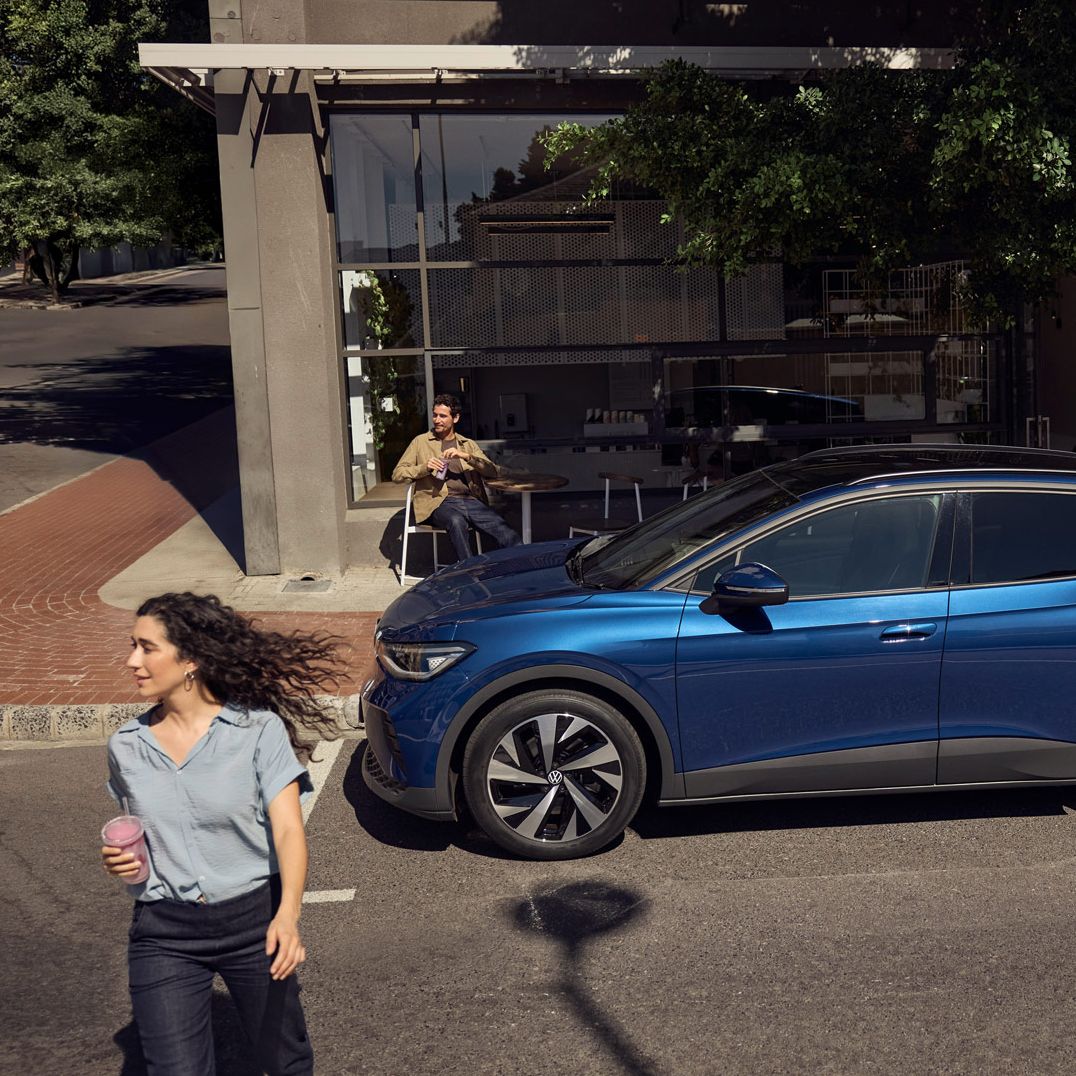 Frau geht mit ihrem Smoothie in der Hand vom geparkten ID.4 über die Straßenseite, ein Mann sitzt dahinter auf einem Stuhl mit Tisch.