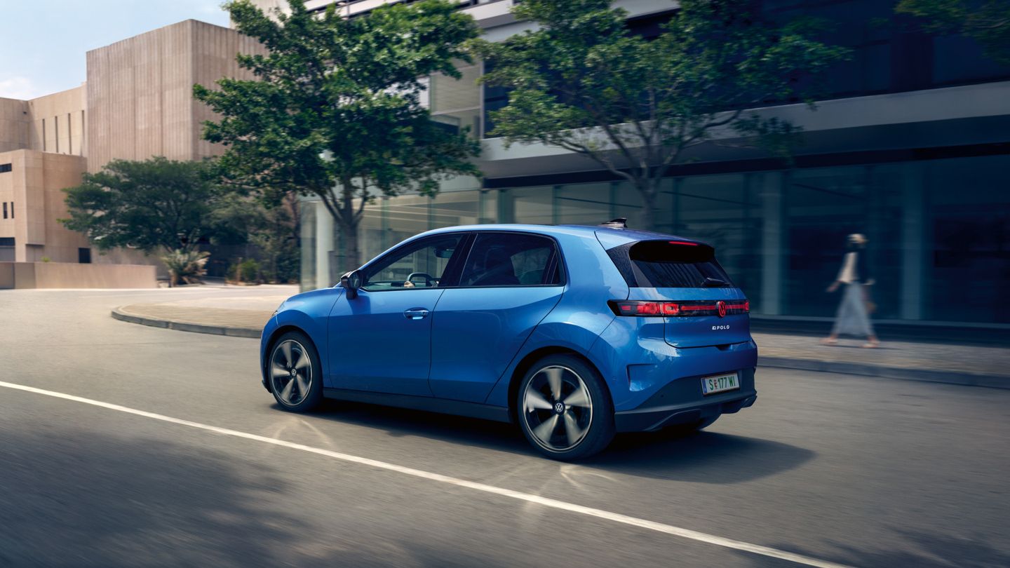Ein blauer VW ID. Polo Style fährt durch eine Stadt mit grünen Bäumen am Straßenrand.
