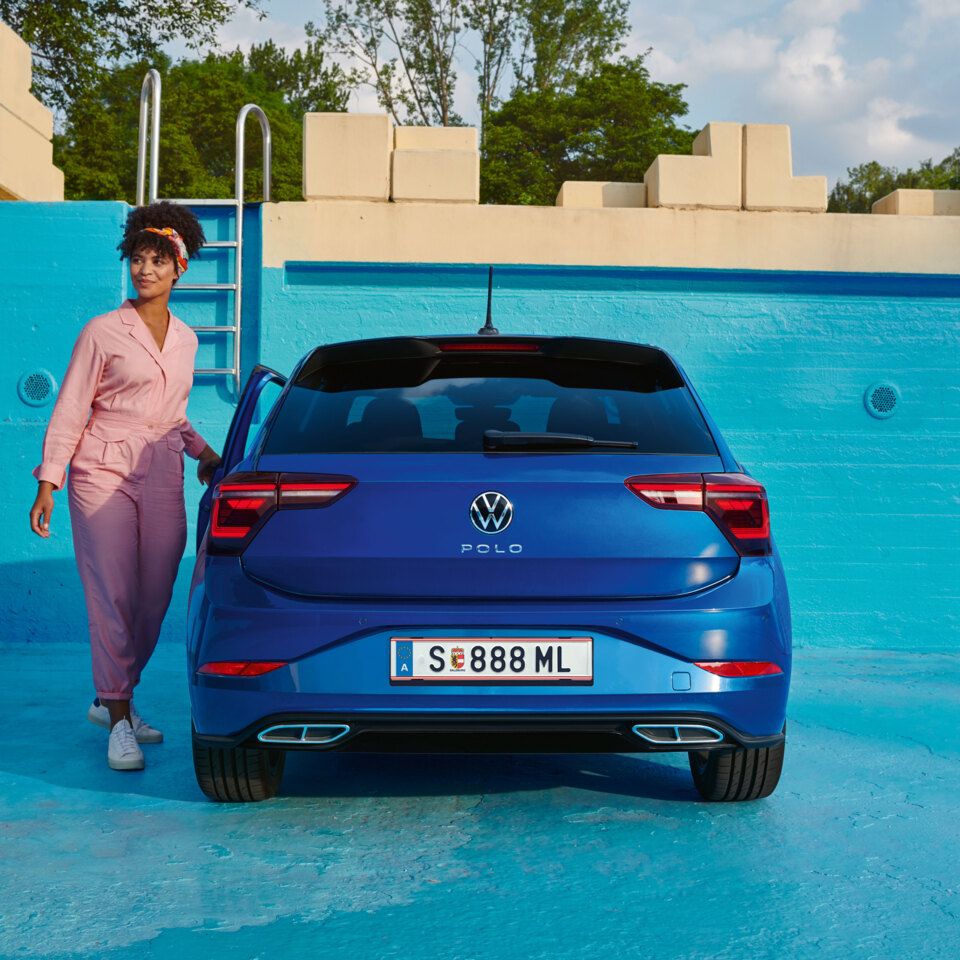 Ein blauer VW Polo steht in einem blau angemalten, leeren Poolbecken, eine junge Frau steigt ein.