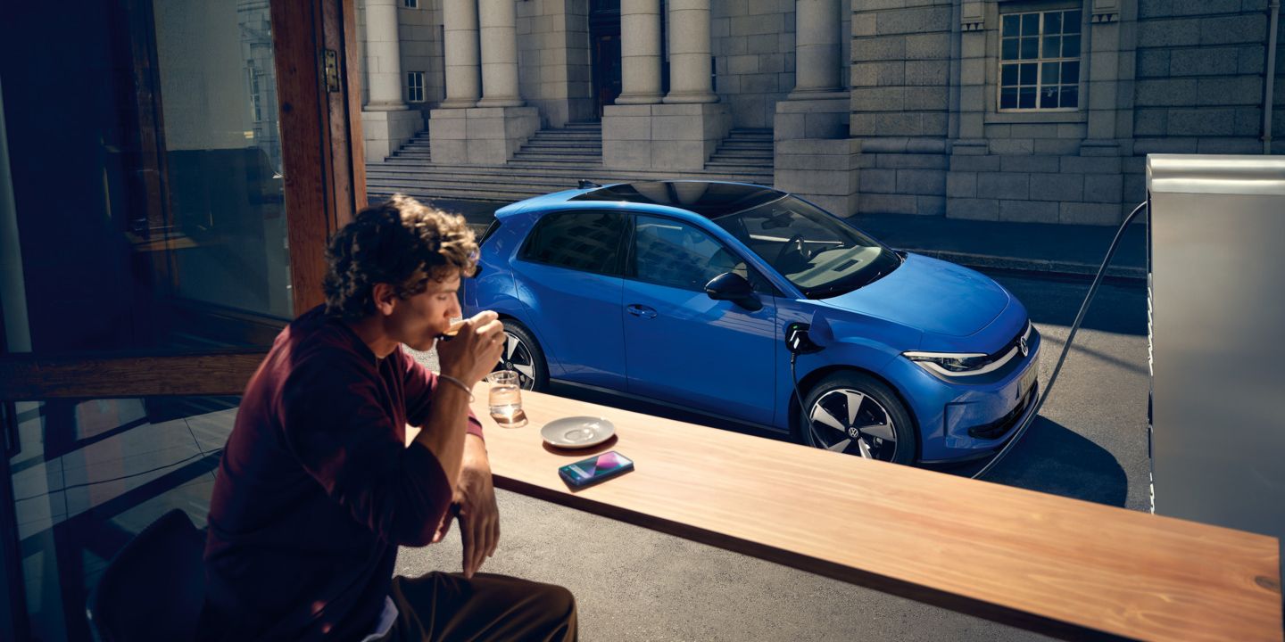 Ein Mann sitzt in einem Café, durch ein offenes Fenster ist der blaue VW ID. Polo Style zu sehen, der an einer Ladestation lädt.