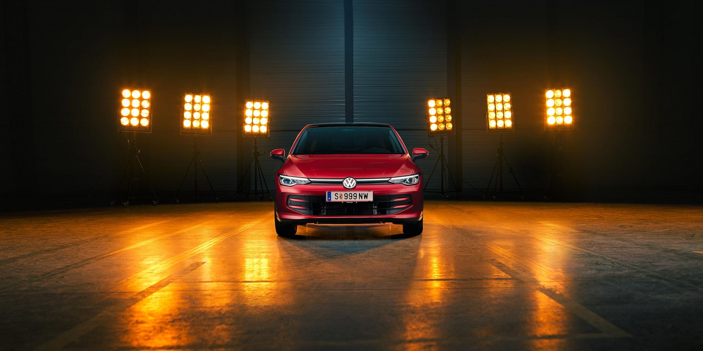Ein roter VW Golf Falco Edition in Frontalansicht steht mittig im Bild in einer leeren Halle, Bühnenlichter strahlen das Fahrzeug von hinten an.