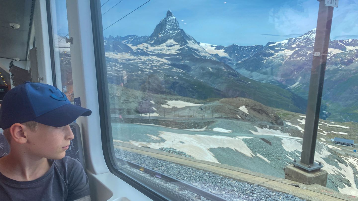 Eine Person sitzt in einem Zug und schaut durch das Fenster auf die schneebedeckten Alpen. Im Hintergrund ist das Matterhorn zu sehen. Der Himmel ist klar und blau.