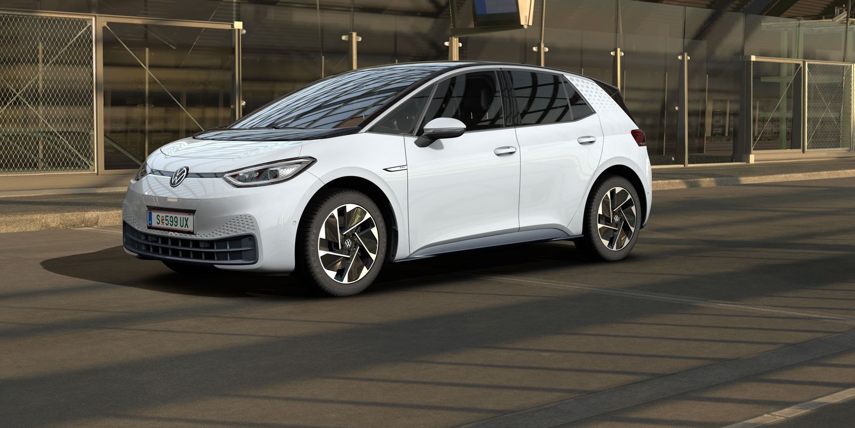 Ein weißer Volkswagen ID.3 steht in einer modernen, teilweise verglasten Tiefgarage. Das Auto ist von der Seite und leicht von vorne zu sehen. Im Hintergrund sind Maschendrahtzäune und Betonwände zu erkennen.
