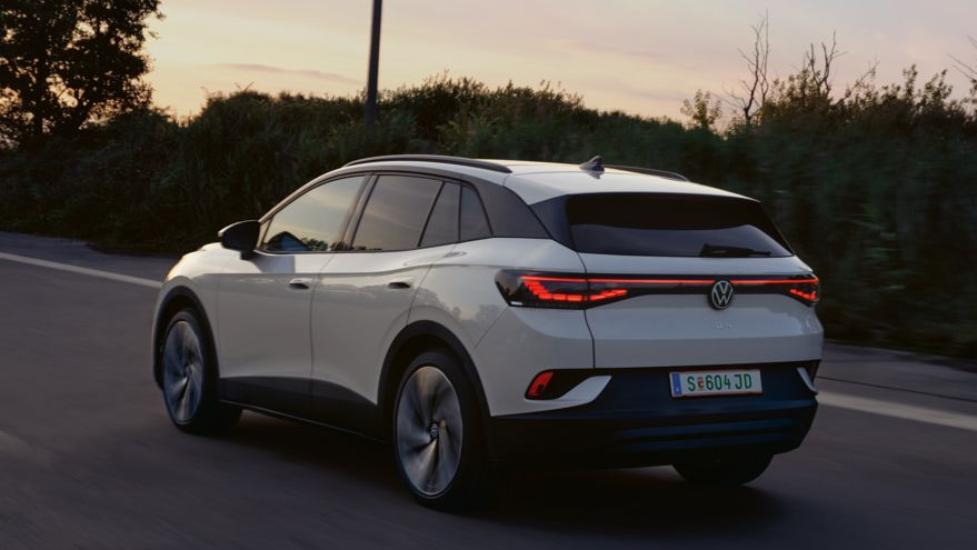 Ein weißer VW ID.4 fährt auf einer Landstraße bei Sonnenuntergang. Das Auto ist von hinten zu sehen. Die Straße ist von Bäumen und Sträuchern gesäumt.