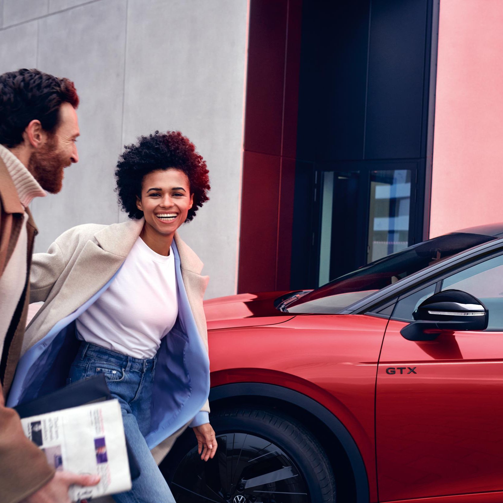 Ein roter VW ID.4 GTX parkt am Straßenrand, ein Mann und eine Frau gehen lachend darauf zu.
