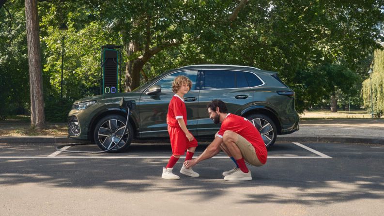 Ein Mann mit seinem Kind in Fußballtrikots steht vor einem VW Tiguan R-Line Plug-In-Hybrid auf einem Parkplatz.