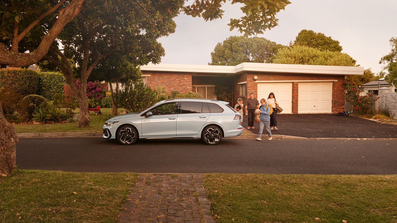 Weißer VW Golf Variant parkt in einer Wohnstraße mit Bäumen. Familie geht mit Gepäck darauf zu.