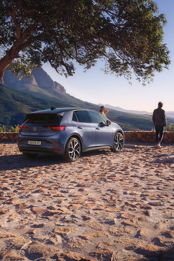 Auf einem Parkplatz in den Bergen parkt ein blauer VW ID.3 Neo, eine Frau und ein Mann genießen die Aussicht