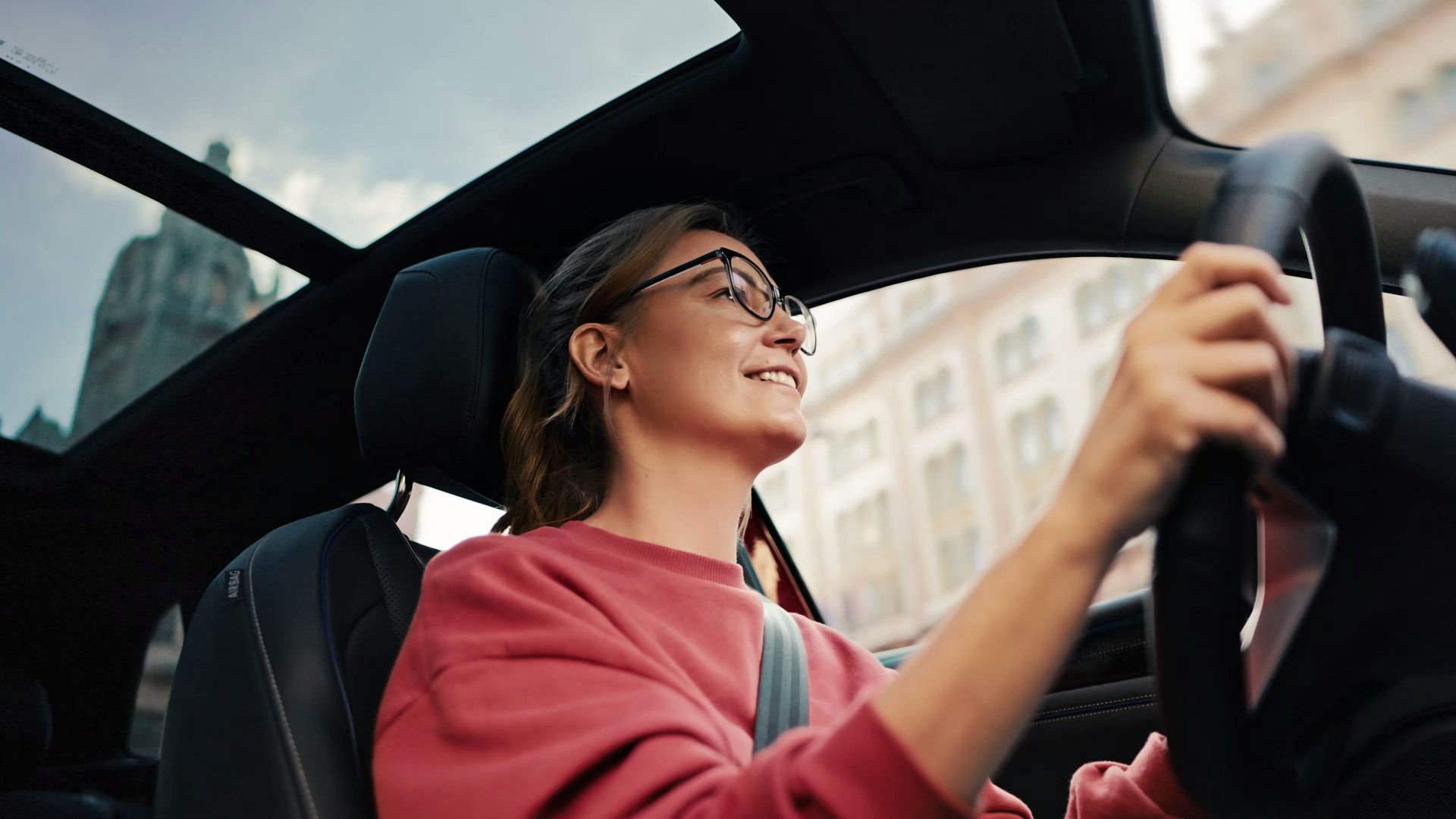 Eine Person in einem roten Pullover sitzt am Steuer eines Autos und fährt durch eine Stadt. Das Gesicht der Person ist unscharf gemacht. Das Auto hat ein großes Schiebedach und im Hintergrund sind Gebäude erkennbar.