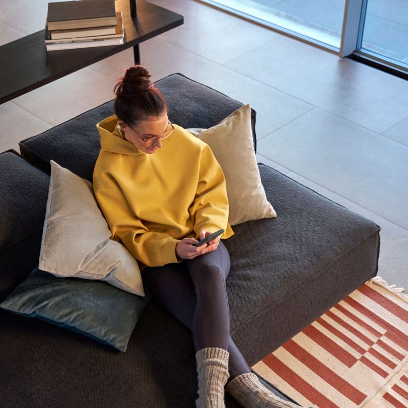 Person sitzt in einem modernen Wohnzimmer auf einem grauen Sofa und hält ein Handy in der Hand. Sie trägt einen gelben Hoodie, schwarze Leggings und graue Socken. Ein Bücherregal voller Bücher steht im Hintergrund.