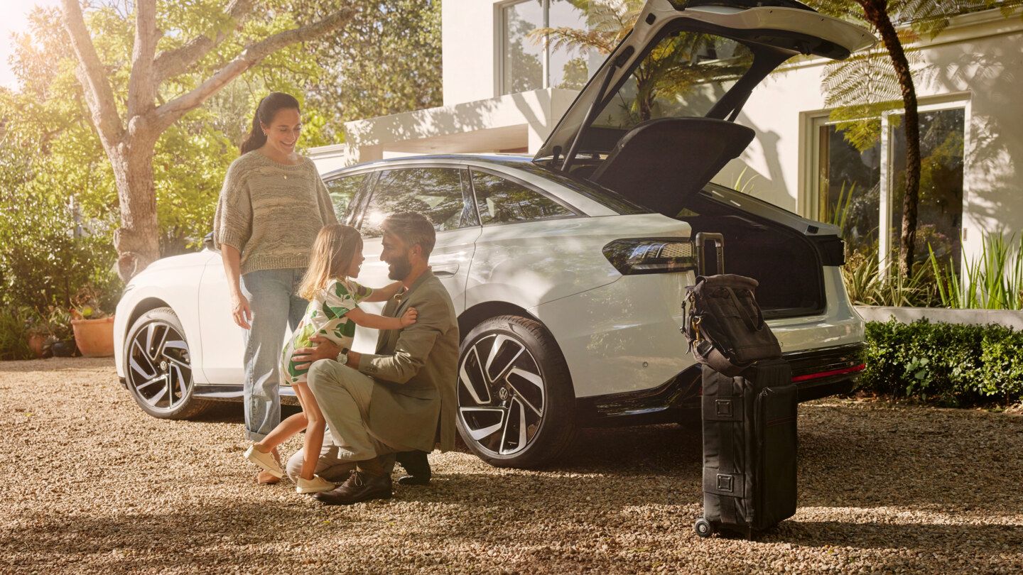 Ein weißer VW ID.7 mit geöffneter Heckklappe steht vor einem Wohnhaus, ein Mann kniet davor und nimmt seine Tochter in den Arm.