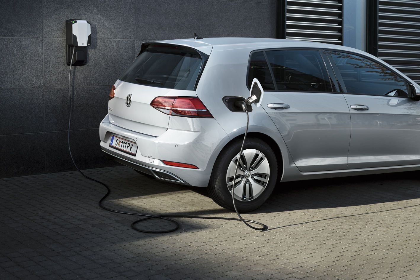 Ein silberner VW Elektroauto e-Golf wird an einer Ladestation aufgeladen. Das Ladekabel ist an die Rückseite des Fahrzeugs angeschlossen. Die Station befindet sich neben einer Wand.