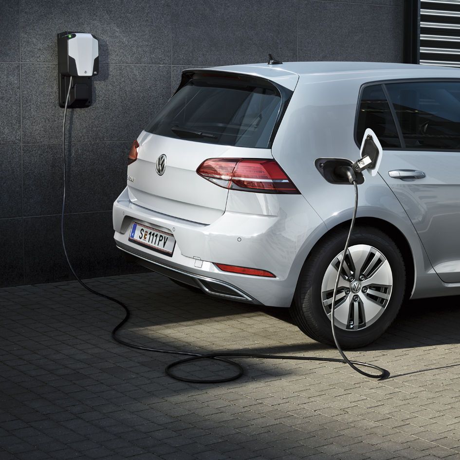 Ein silberner VW Elektroauto e-Golf wird an einer Ladestation aufgeladen. Das Ladekabel ist an die Rückseite des Fahrzeugs angeschlossen. Die Station befindet sich neben einer Wand.
