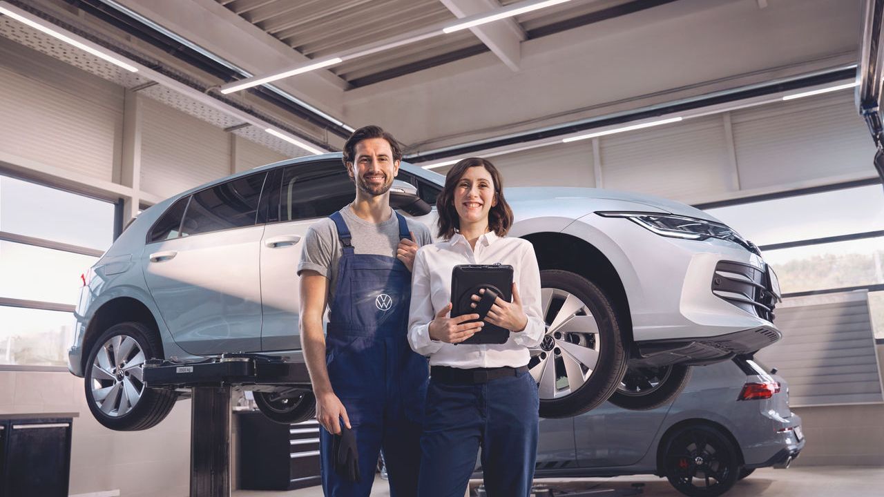 Zwei Volkswagen Mitarbeiter:innen lachen in die Kamera. Im Hintergrund ein Volkswagen Golf auf einer Hebebühne.
