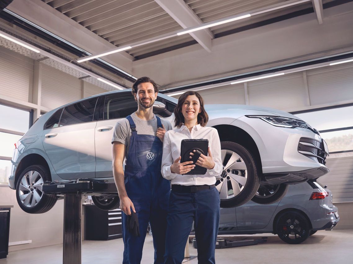 Zwei Volkswagen Mitarbeiter:innen lachen in die Kamera. Im Hintergrund ein Volkswagen Golf auf einer Hebebühne.