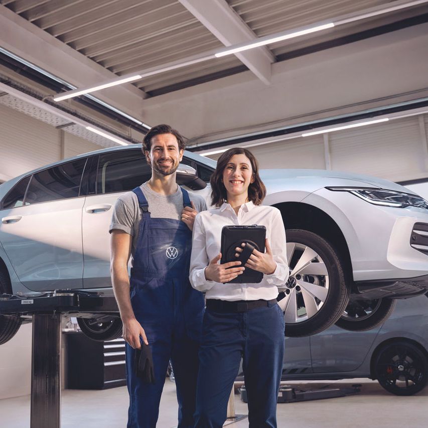 Zwei Volkswagen Mitarbeiter:innen lachen in die Kamera. Im Hintergrund ein Volkswagen Golf auf einer Hebebühne.