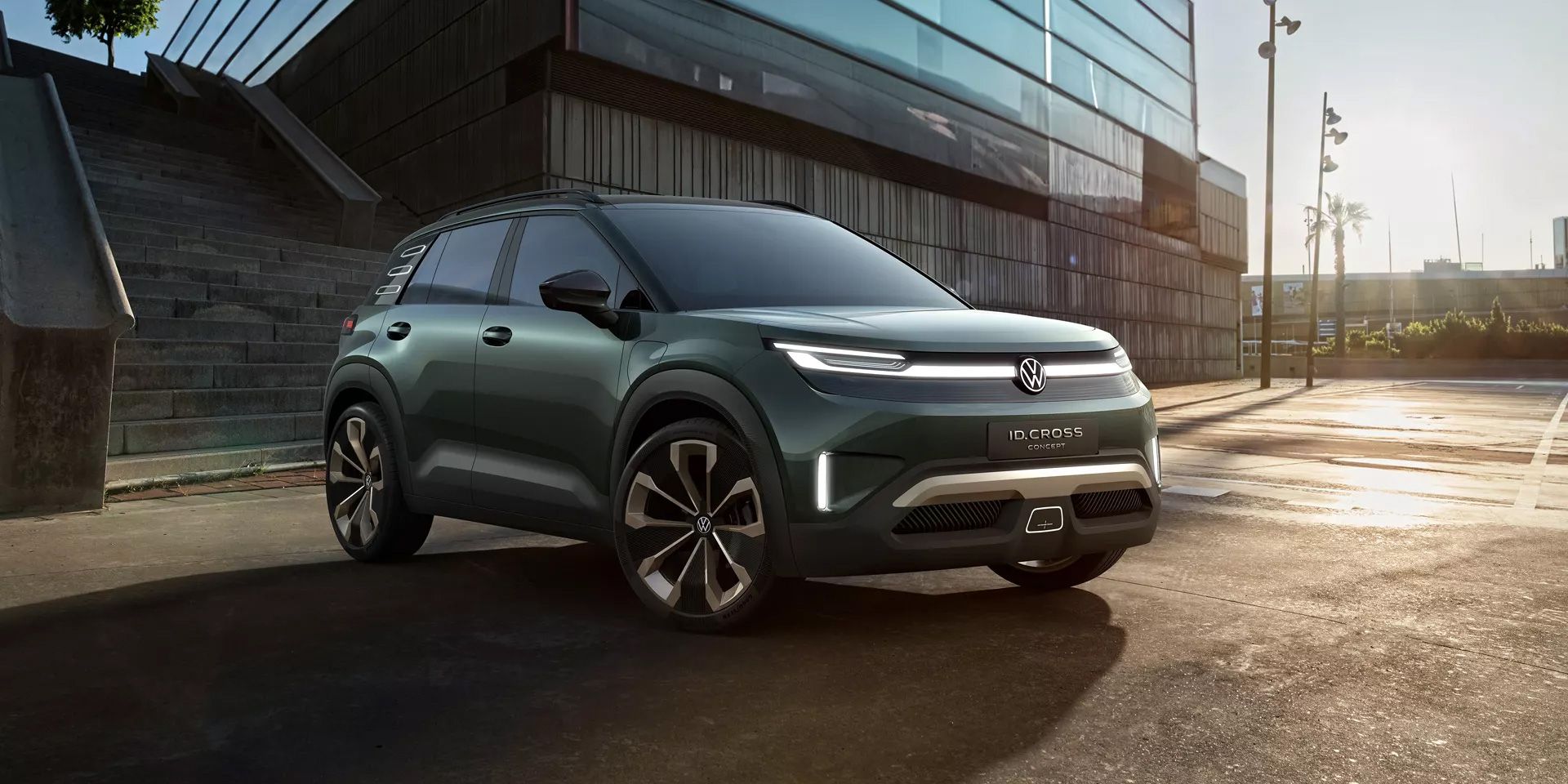 Ein dunkelgrünes Volkswagen ID. Cross steht auf einem Parkplatz vor einem modernen Gebäude mit Glasfassade. Der SUV hat futuristische Scheinwerfer und stylische Felgen. Im Hintergrund sind Treppenstufen und ein paar Palmen sichtbar.