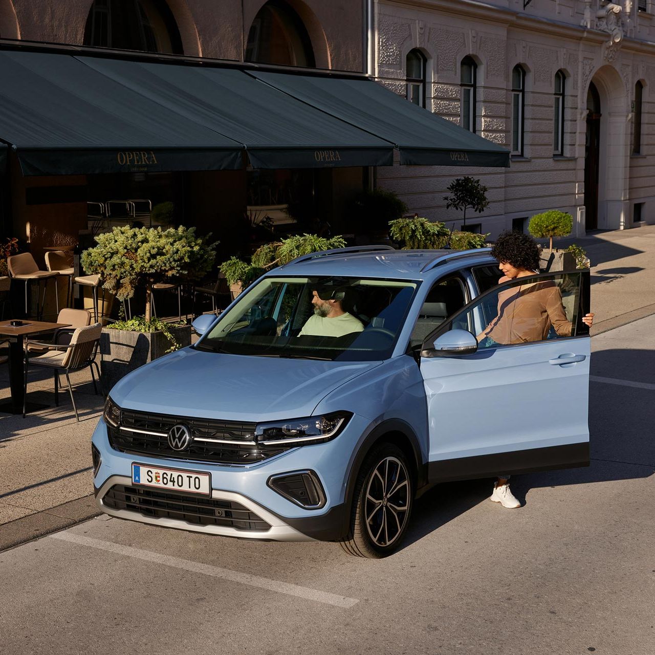 Eine Frau steigt in einen blauen VW T-Cross, der vor einem Straßencafé parkt.