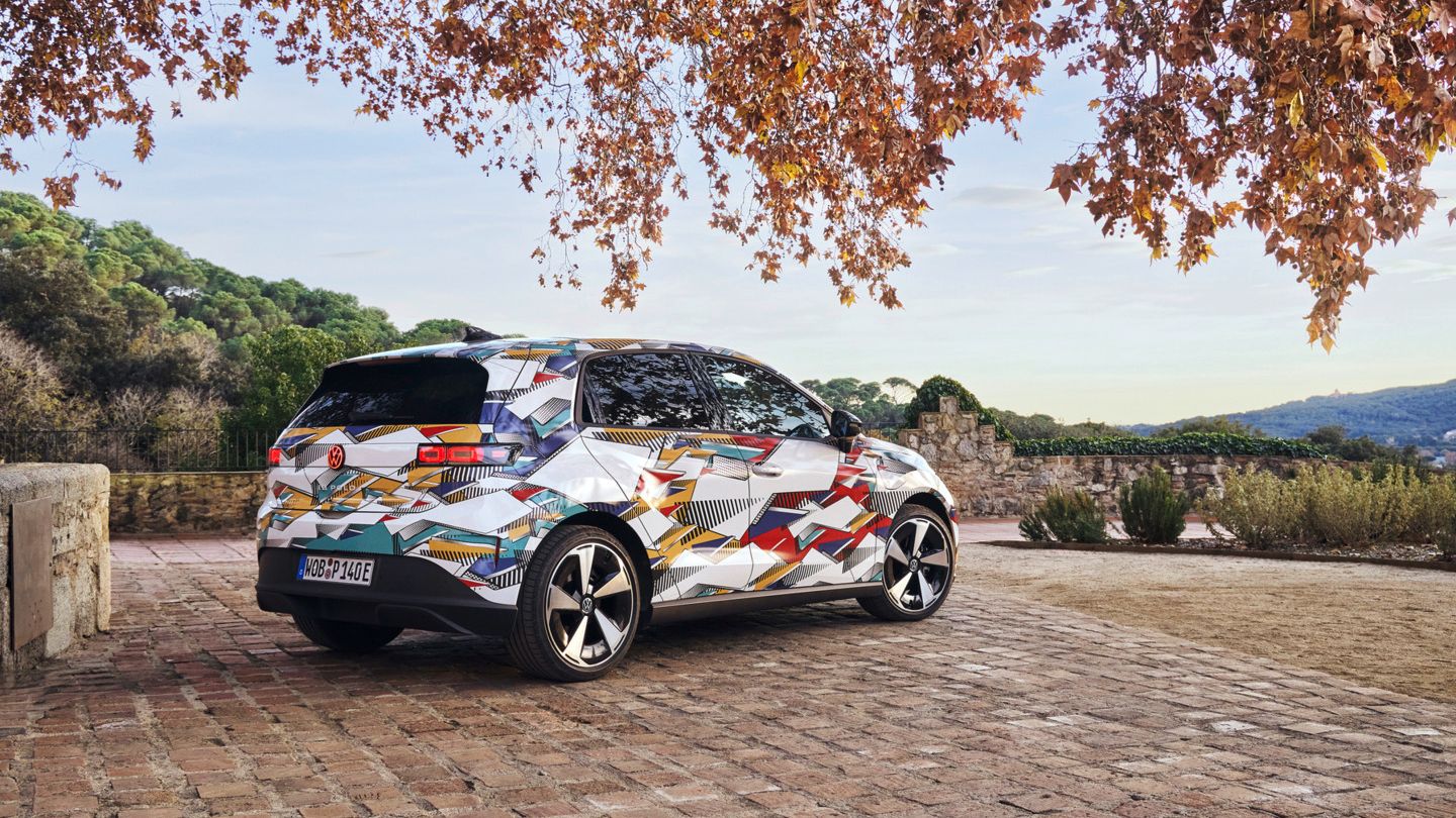 Ein ID. Polo mit bunter geometrischer Folierung steht auf einem gepflasterten Hof. Im Hintergrund sind Hügel mit Bäumen sichtbar, und der Himmel ist teilweise bewölkt. Ein Baum mit Herbstblättern ragt von oben in das Bild hinein.