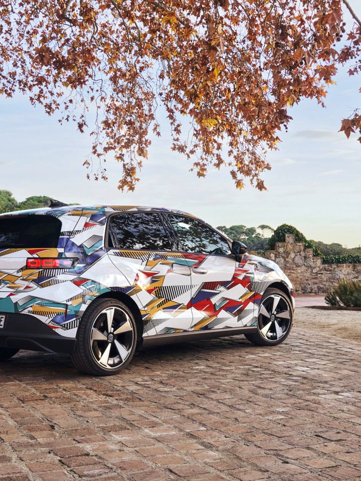 Ein ID. Polo mit bunter geometrischer Folierung steht auf einem gepflasterten Hof. Im Hintergrund sind Hügel mit Bäumen sichtbar, und der Himmel ist teilweise bewölkt. Ein Baum mit Herbstblättern ragt von oben in das Bild hinein.