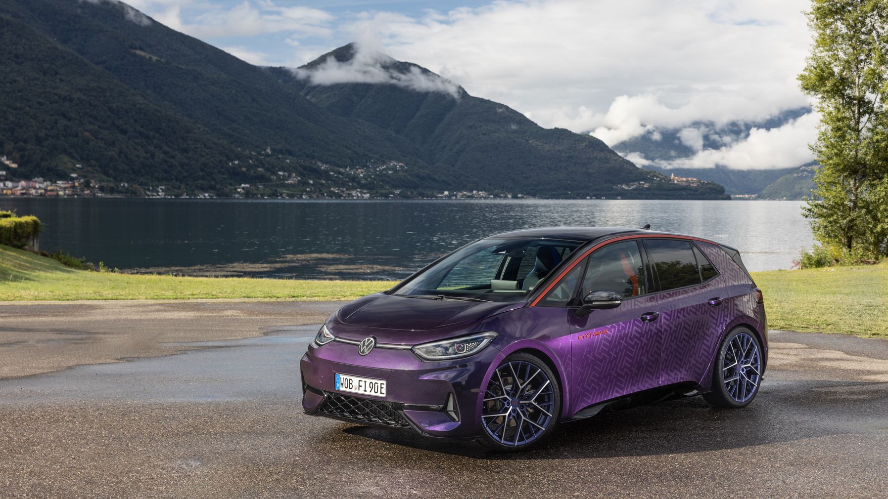 Ein lila Volkswagen-Elektroauto steht auf einem Parkplatz vor einem See und von Bergen umgeben. Der Himmel ist teilweise bewölkt. Das Auto hat ein modernes Design mit blauen Felgen.