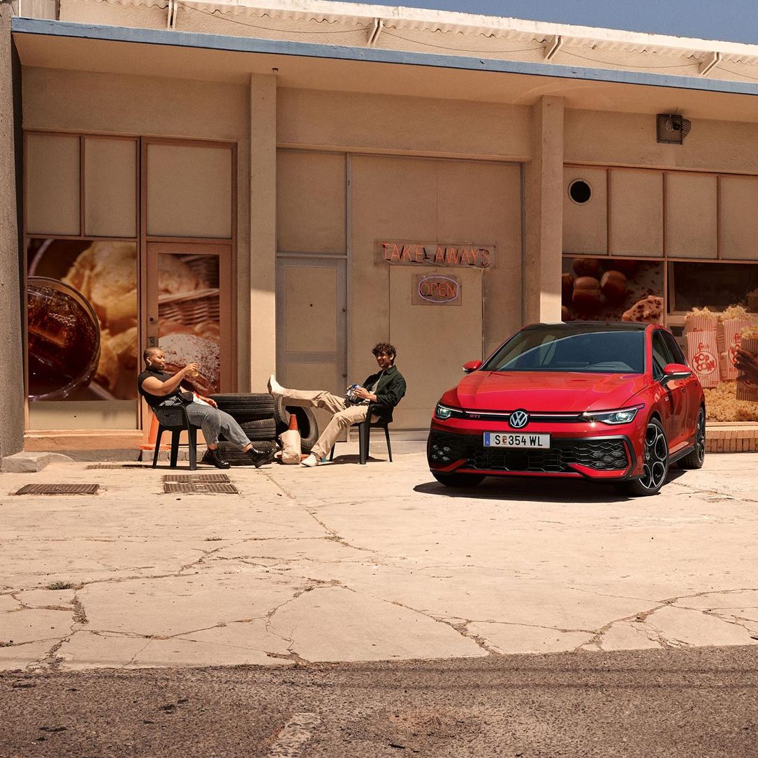 Zwei junge Männer sitzen vor einem Geschäft neben einem roten VW Golf GTI.