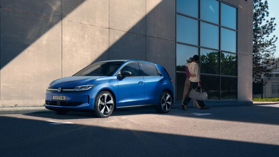 Ein blauer ID. Polo parkt vor einem rechteckigem Haus mit einer großen Fensterfront. Eine Frau und ein Mann mit einer Tasche in der Hand bewegen sich vom Auto weg.